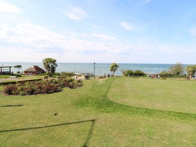 A park with grass, flower beds, benches, palm trees, and a sea view at The Burleigh in Hunstanton