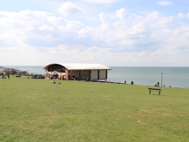 A large grassy area with people sitting and walking near a pavilion building by the sea at The Burleigh in Hunstanton