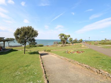 A paved pathway through a grassy park with trees and flower beds by the sea at The Burleigh in Hunstanton