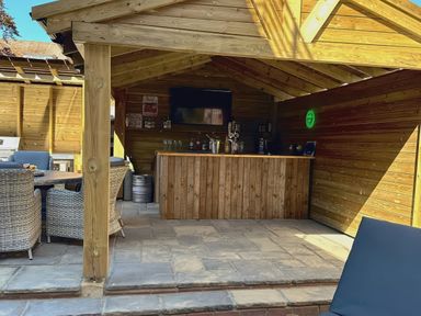 An outdoor wooden bar area with a counter TV chairs and a table under a wooden roof at The Burleigh in Hunstanton