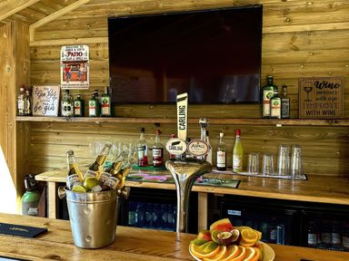 A wooden bar with bottles, glasses, a fruit plate, a bucket with beers, signs, and a large TV mounted on the wall at The Burleigh in Hunstanton