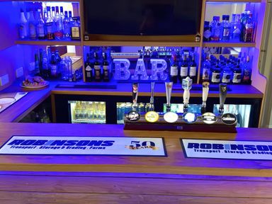 A bar counter with various spirits bottles a lit bar sign and six beer taps with a refrigerator stocked with drinks behind the counter at The Burleigh in Hunstanton