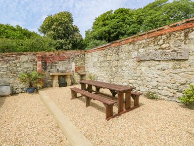 An outdoor seating area with a wooden picnic table and benches on a gravel surface next to a stone wall with a sign that says the Lookout and a built-in barbecue