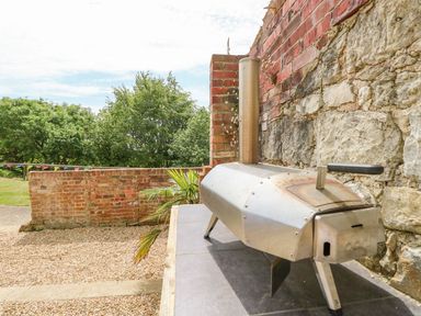 A small metal outdoor pizza oven on a tiled surface next to a brick and stone wall with trees and a brick fence in the background at Moonlight in Ventnor