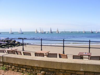 A beach with multiple sailboats on the water and wooden chairs and tables along a promenade at Moonlight in Ventnor