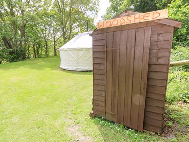 A brown woodshed with a wooden sign next to a white tent on grass with trees in the background at Stardust in Ventnor