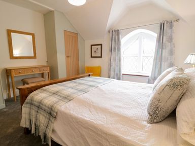 A bedroom with a wooden bed a plaid throw pillows a wooden desk with a mirror a yellow chair and arched window with curtains at Dwynant in Bontddu near Dolgellau