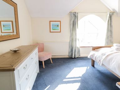 A bedroom with a wooden bed, white dresser, pink chair, and an arched window with curtains at Dwynant Bontddu near Dolgellau