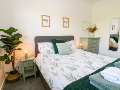 A bedroom with a bed with botanical print bedding green pillows green nightstand with lamp a green chest of drawers with a vase and mirror and a potted plant at Gilfach Bach in Dolgellau
