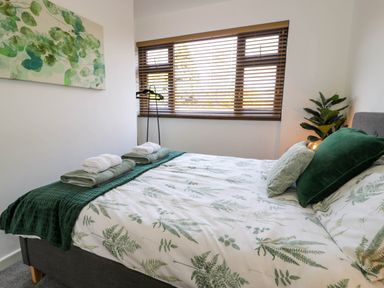 A bedroom with a bed made with leaf patterned bedding green pillows and towels on the bed and a window with wooden blinds at Gilfach Bach in Dolgellau
