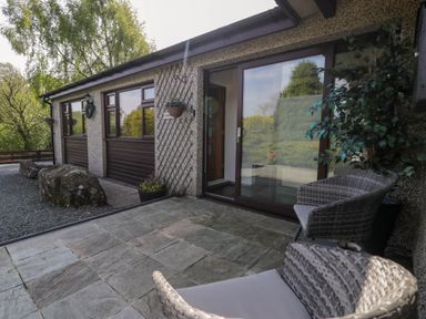 An outdoor patio with stone tiles and wicker chairs next to a building with large windows and gravel pathway at Gilfach Bach in Dolgellau