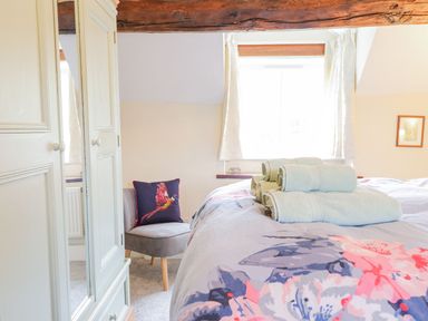 A bedroom with a bed covered with floral bedding and folded towels next to a window with white curtains and a gray chair with a bird pillow at Ty Glan Yr Afon in Bontddu near Dolgellau