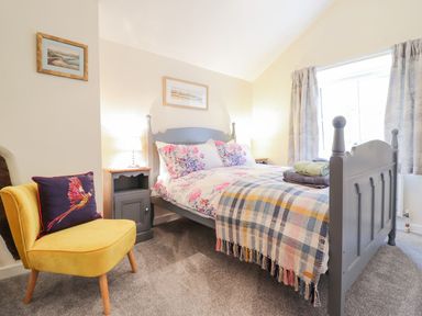 A bedroom with a grey bed and floral bedding a yellow chair with a bird cushion and bedside tables with lamps at Ty Glan Yr Afon in Bontddu near Dolgellau