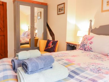 A bedroom with a bed covered in floral bedding and folded towels on it a yellow chair with a cushion a grey wardrobe with mirrored doors and a wooden door at Ty Glan Yr Afon in Bontddu near Dolgellau