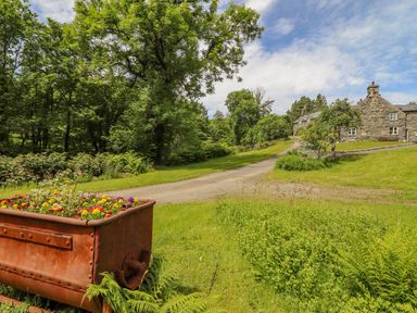 A garden with a rusty metal planter filled with flowers a gravel driveway and stone houses surrounded by trees at Ty Glan Yr Afon in Bontddu near Dolgellau