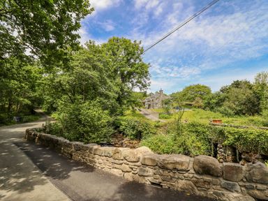 A stone wall along a road with trees and a stone house in the background at Ty Glan Yr Afon in Bontddu near Dolgellau