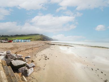 A pebble and sandy beach with small blue and white beach huts and a grassy hill under a cloudy sky