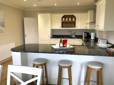 A kitchen with three wooden stools at a black countertop and cream cabinets at Pen Y Frith Bungalow in Prestatyn
