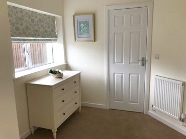 A room with a white dresser under a window with floral blinds a framed picture on the wall a white door and a radiator at Pen Y Frith Bungalow in Prestatyn