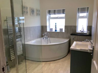A bathroom with a corner bathtub a sink two windows and a towel rack at Pen Y Frith Bungalow in Prestatyn