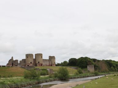 A stone castle ruin on a hill beside a river with trees and sheep in a grassy field near Pen Y Frith Bungalow in Prestatyn