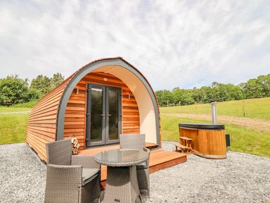 A wooden pod with double glass doors a small patio with a round table and two chairs and a hot tub outside on gravel with grass and trees behind at Briallen in Bontddu near Dolgellau