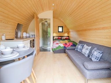 An interior view of a wooden pod with a bed floral bedding a gray sofa with cushions a small kitchen and a dining table at Briallen in Bontddu near Dolgellau