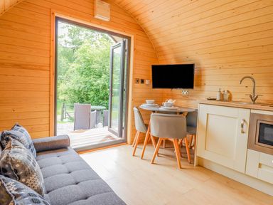 A small wooden cabin interior with a gray sofa a round dining table set with dishes four chairs a wall-mounted TV kitchenette and an open door leading to outdoor seating at Briallen in Bontddu near Dolgellau