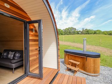 A wooden pod with an open door showing a sofa inside and an outdoor hot tub in a grassy area at Briallen in Bontddu near Dolgellau