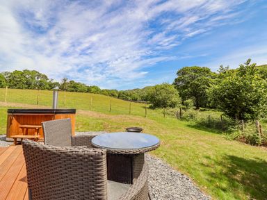 An outdoor seating area with a round table and two wicker chairs beside a wooden hot tub and grassy field at Briallen in Bontddu near Dolgellau