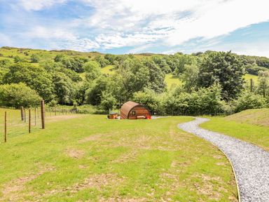 A grassy field with a curved gravel path leading to a small wooden pod surrounded by trees and hills at Briallen in Bontddu near Dolgellau