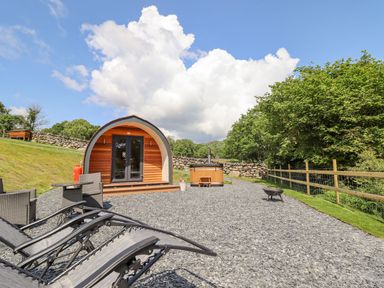 A wooden pod with glass doors and outdoor chairs and sun loungers on a gravel area with trees and a stone wall at Eirlys in Bontddu near Dolgellau