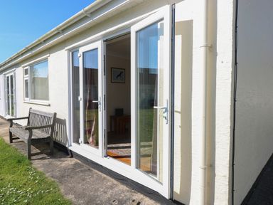 A building exterior with open glass doors next to a wooden bench and grass at Kites - 52 Gower Holiday Village in Scurlage near Port Eynon and Rhossili