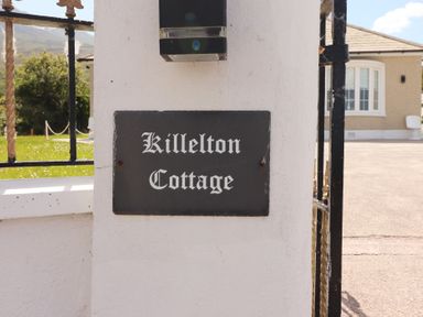 A white wall with a black rectangular sign reading Killelton Cottage next to a gate and a house in the background at Killelton Cottage in Camp County Kerry