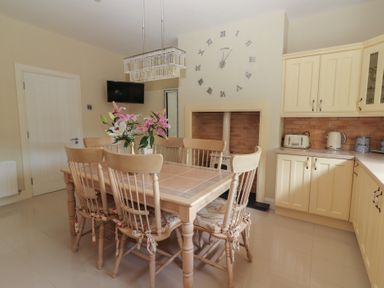 A kitchen dining area with a wooden table and six chairs a vase of flowers a wall clock a small tv and cream cabinets at Killelton Cottage Camp County Kerry