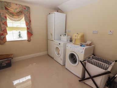 A laundry room with a washing machine, dryer, refrigerator, laundry basket, and a window with floral curtains at Killelton Cottage in Camp County Kerry