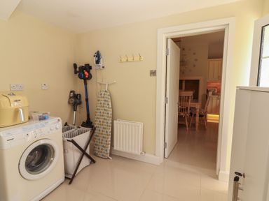 A utility room with a washing machine ironing board laundry basket and vacuum cleaners next to a doorway leading to a kitchen with wooden chairs at Killelton Cottage in Camp County Kerry