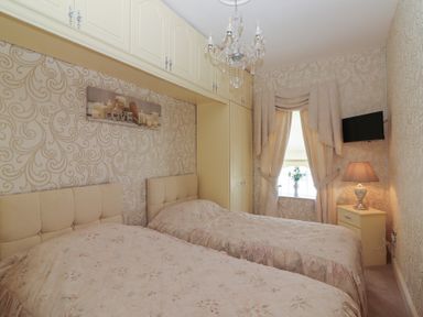 A bedroom with two single beds a chandelier built in cupboards curtains bedside table with lamp and a wall mounted television at Killelton Cottage in Camp County Kerry