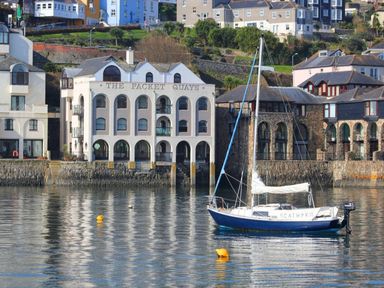 A sailboat on water in front of buildings including one labeled The Packet Quays at Tobys Quay in Falmouth