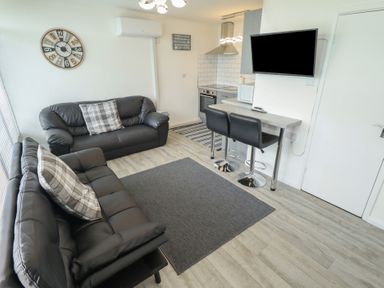 A living room with two black sofas a wall clock and a kitchen area with bar stools and a wall-mounted tv at 99 Cherry Park in Chapel St Leonards