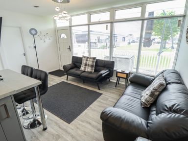 A living room with two black leather sofas a small wooden side table bar stools and large windows at 99 Cherry Park in Chapel St Leonards