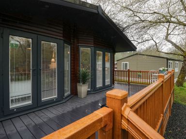 An outdoor deck with wooden railing and black wood flooring outside a cabin with glass doors and a potted plant at Robins Lodge White Cross Bay near Troutbeck Bridge