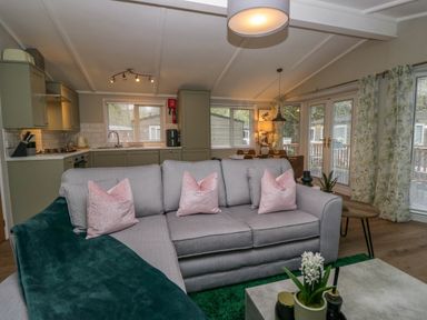 An open plan living area with a grey sofa pink cushions and green throw a kitchen and dining area with wooden table glass doors and floral curtains at Robins Lodge in White Cross Bay near Troutbeck Bridge