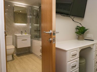 A bathroom with a toilet sink bathtub and mirror visible through an open door next to a desk with a plant and TV on the wall at Robins Lodge White Cross Bay near Troutbeck Bridge