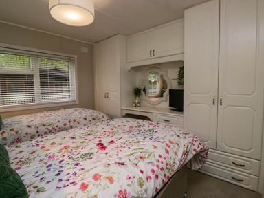 A bedroom with two beds with floral bedding a window with blinds and built in white wardrobes and a desk with a mirror at Robins Lodge White Cross Bay near Troutbeck Bridge