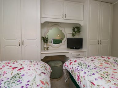 A bedroom with two beds with floral bedding a white built-in wardrobe a wall mirror a small TV and a green cushioned stool at Robins Lodge in White Cross Bay near Troutbeck Bridge