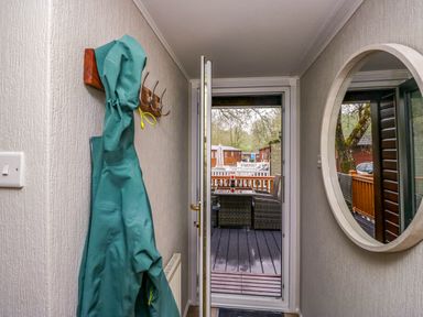 A hallway with a green coat hanging on hooks and a round mirror on the wall showing an outdoor deck with wicker furniture at Robins Lodge in White Cross Bay near Troutbeck Bridge