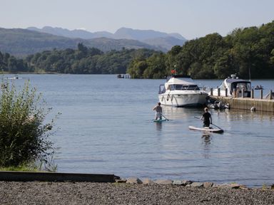 Two people paddleboarding on a lake near boats docked at a pier with forested hills and mountains in the background at Robins Lodge in White Cross Bay near Troutbeck Bridge