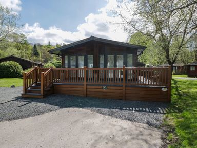A wooden lodge with a front deck and steps surrounded by trees and grass at Robins Lodge White Cross Bay near Troutbeck Bridge