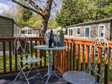 A wooden deck with a metal table and two chairs with wine glasses and a flower pot at Robins Lodge White Cross Bay near Troutbeck Bridge